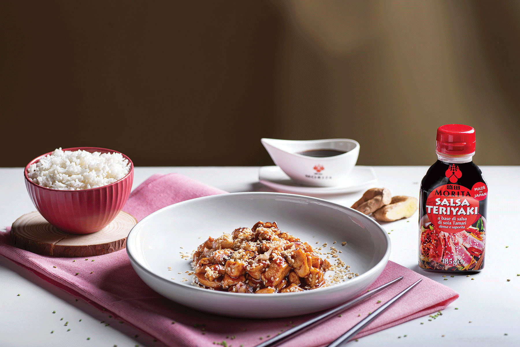 Un piatto di bocconcini di pollo in salsa Teriyaki accanto, da un lato, a una ciotola di riso, e dall'altro, alla bottiglietta di salsa Teriyaki Morita.