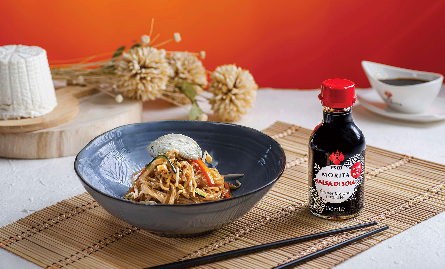 Una ciotolo di spaghetti di riso con verdure e quenelle di ricotta alle erbette accanto alla bottiglietta di salsa di soia tradizionale Morita.